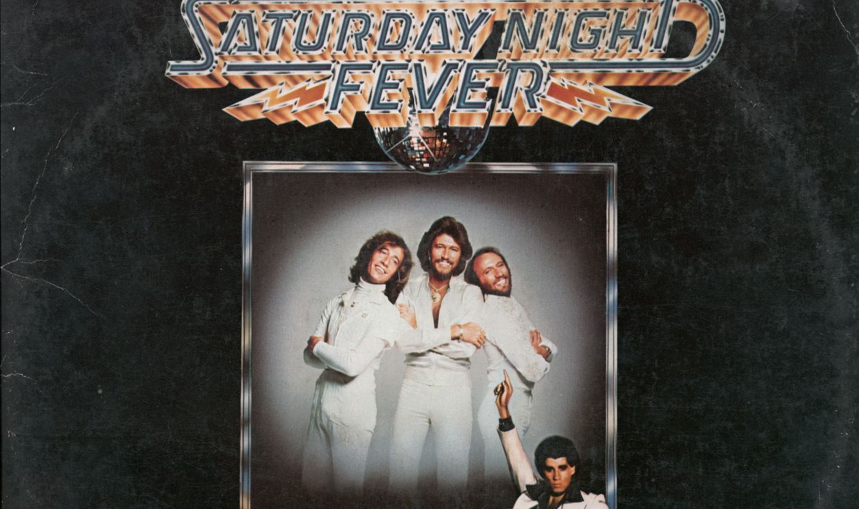 Getty Images - 78263412 - View of the cover of the soundtrack album from the film 'Saturday Night Fever,' 1977. Published by RSO Records, the album's jacket features a large, framed portrait of Australian pop group the Bee Gees as they overlook a photo of American actor John Travolta as he strikes a pose on a colored disco dance floor.