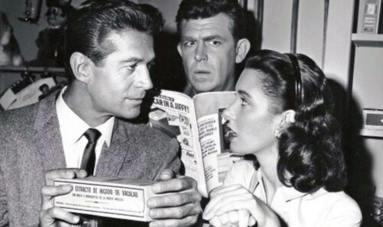 Photo of Andy Griffith, Elinor Donahue and George Nader from the television program The Andy Griffith Show.  Andy can't keep his mind on his magazine as he's trying to hear every word Ellie the pharmacist and Mayberry's new doctor (Nader) are saying.