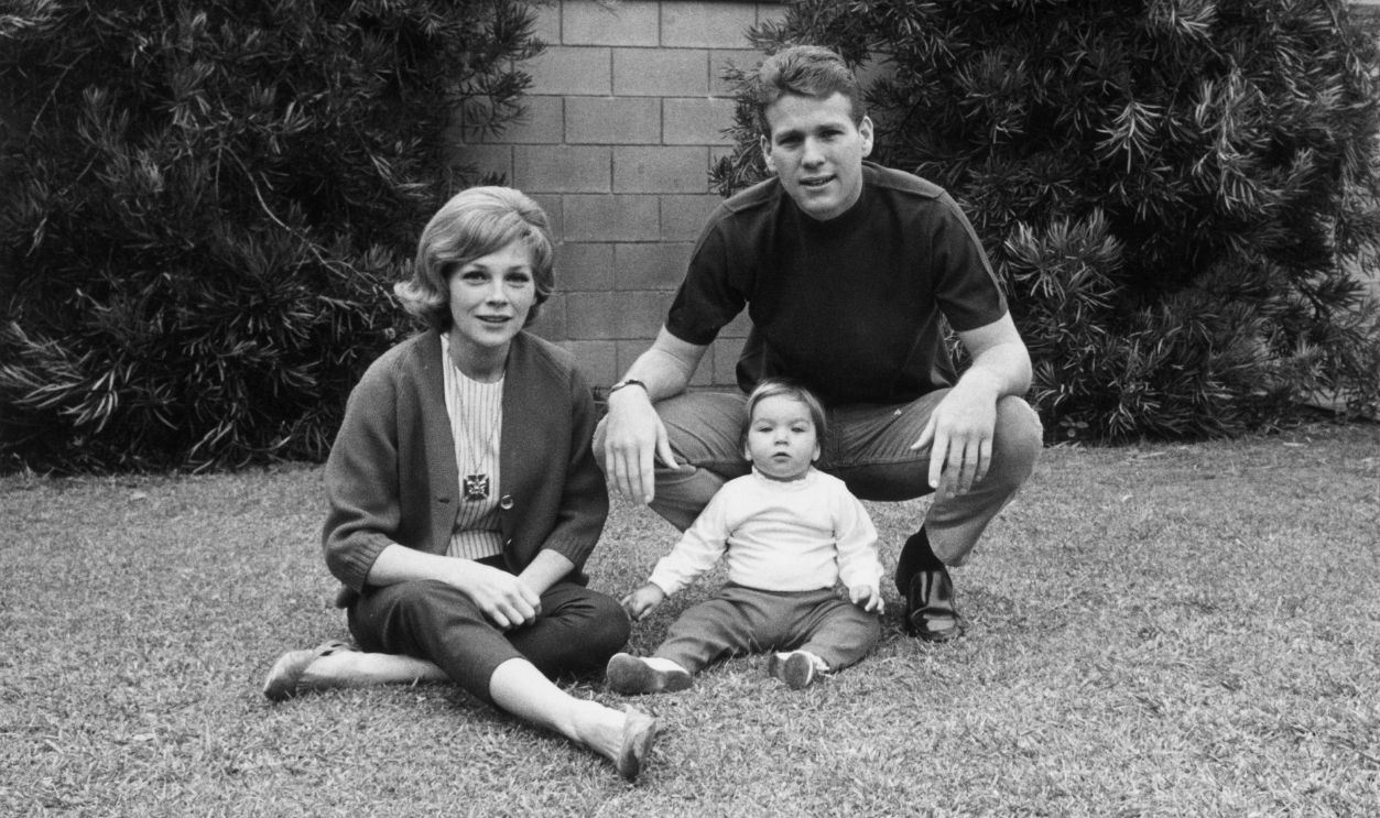 American actor Ryan O'Neal with his wife, actor Joanna Moore, and daughter, Tatum, sitting on grass in front of a stone wall.