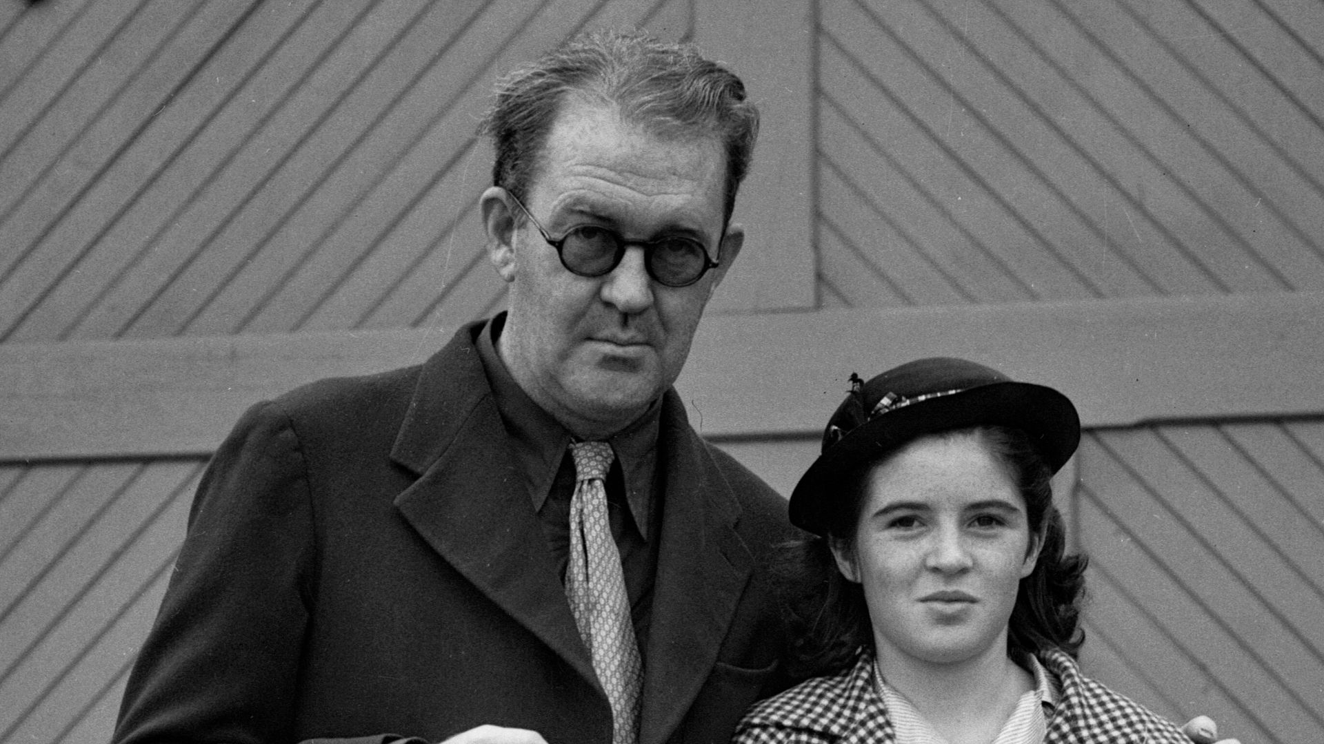John Ford and his daughter Barbara Ford after returning from Hawaii. They are outside, and John holds a pipe while Barbara holds a ukulele and a purse. Text from newspaper caption: John Ford, Fox director, returned on the Matson liner Malolo with his fami