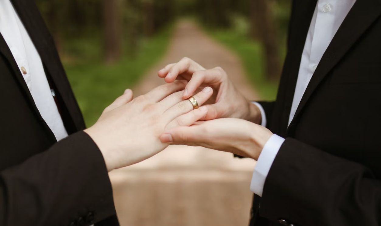Two men in suits exchange wedding rings outdoors