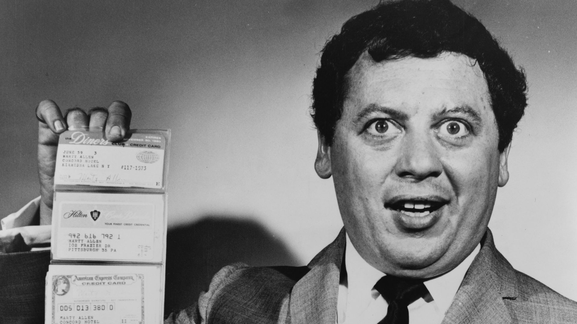 Marty Allen, American comedian, holding wallet with six cards extended in accordion fashion. Visible from top to bottom are