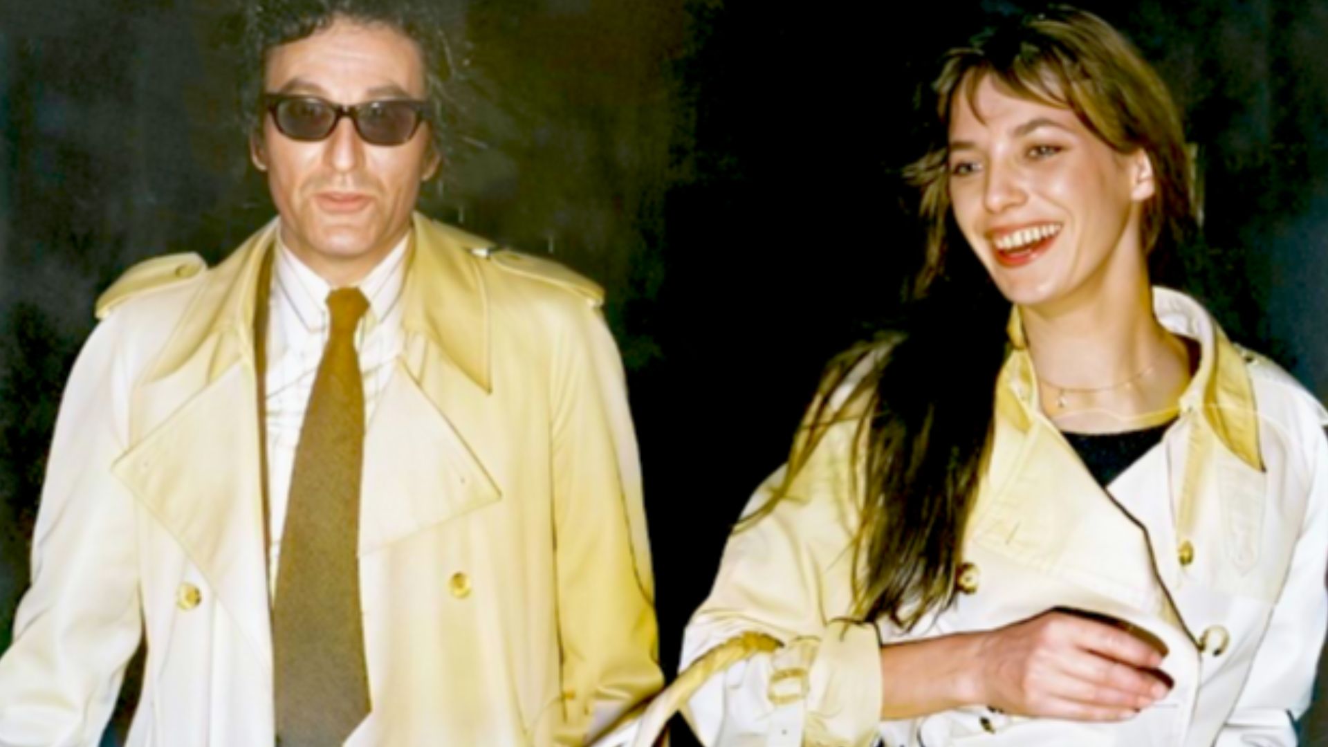Un couple, la nuit, photographiés au flash. Lui, à gauche, est un homme d'une quarantaine d'années, face caméra, cheveux bruns ondulés et portant des lunettes noires, un pardessus mastic et une chemise blanche avec cravate grise. Elle, à droite, est une j