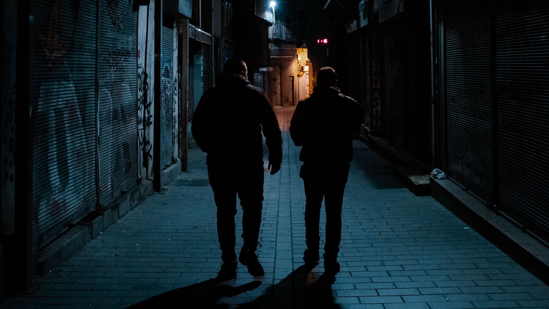 Two men silhouette walking in a dark urban alley at night with street lamps.