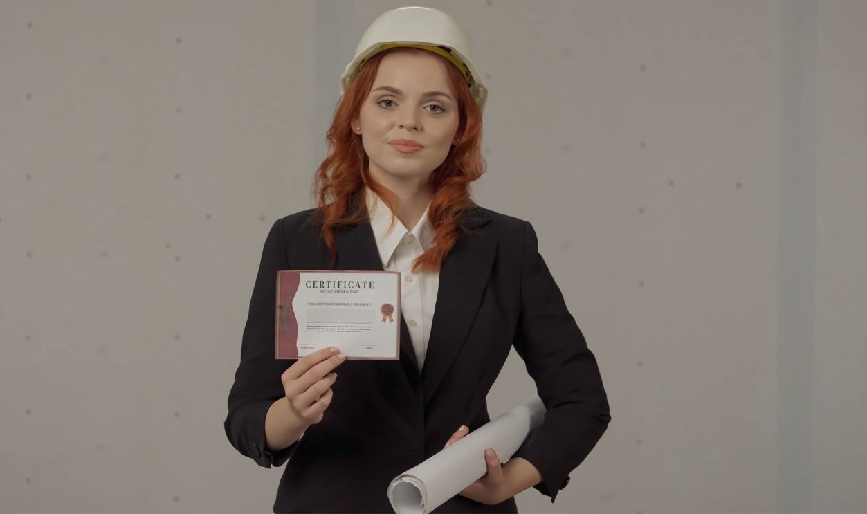 Shutterstock - 2354684633 - A woman architect holds a certificate in her hand, confirming her high qualification. Portrait of a business woman in a construction helmet, with blueprints and a certificate in her hands.