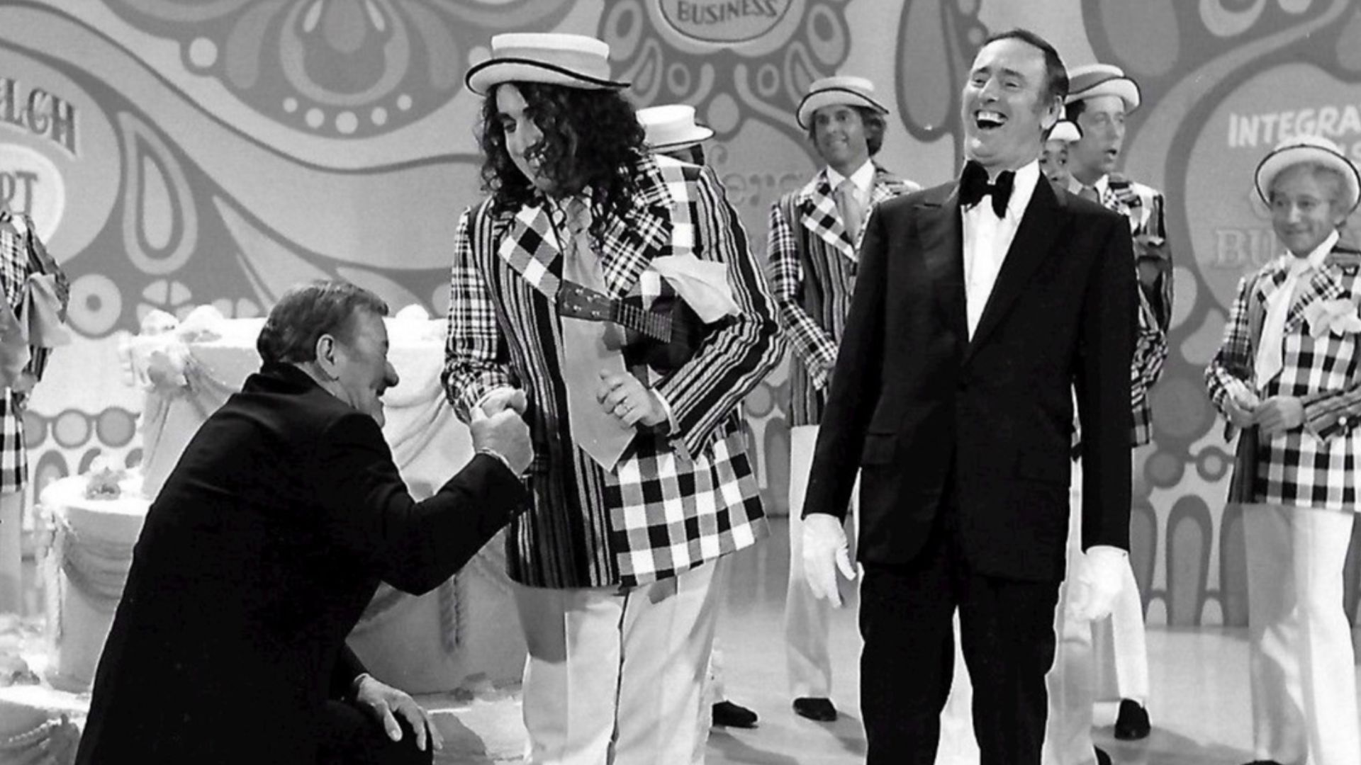 Publicity photo from the 100th episode of the television program Rowan & Martin's Laugh-In.  Guest stars are John Wayne and Tiny Tim. Host Dick Martin is pictured, along with past and present cast members from the show.  Visible in the background are Ruth