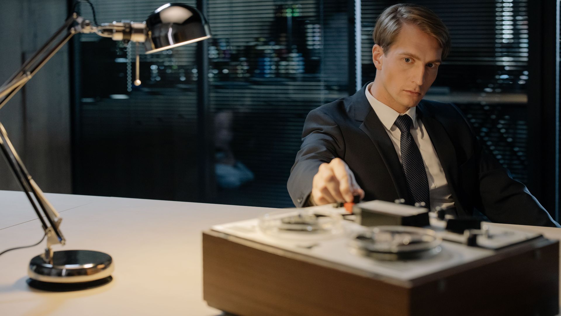 A detective in a suit uses a tape recorder under a desk lamp, evoking vintage investigation themes.