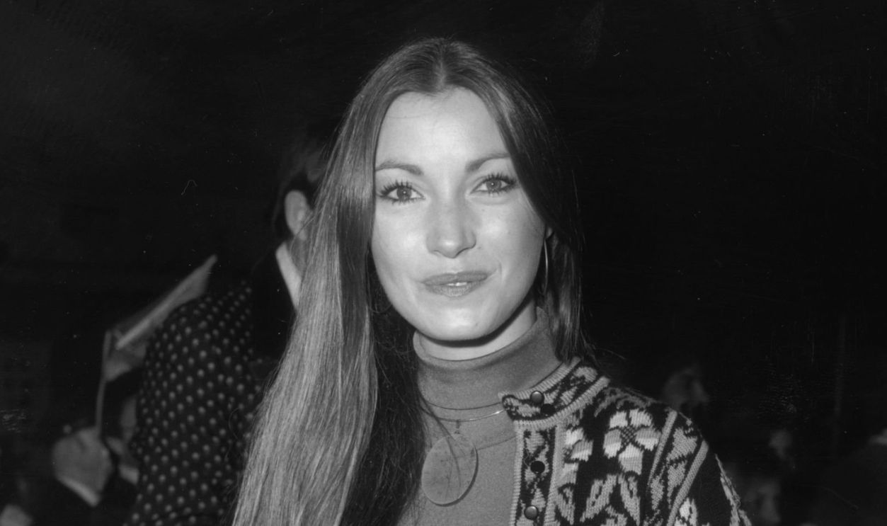 11th November 1975: British-born actor Jane Seymour smiles while standing with her hands behind her back at a block party for the Hollywood premiere of director Susan Winslow's film 'All This and World War II,' Hollywood, California. Seymour wears a knit cardigan and matching skirt with a turtleneck sweater.