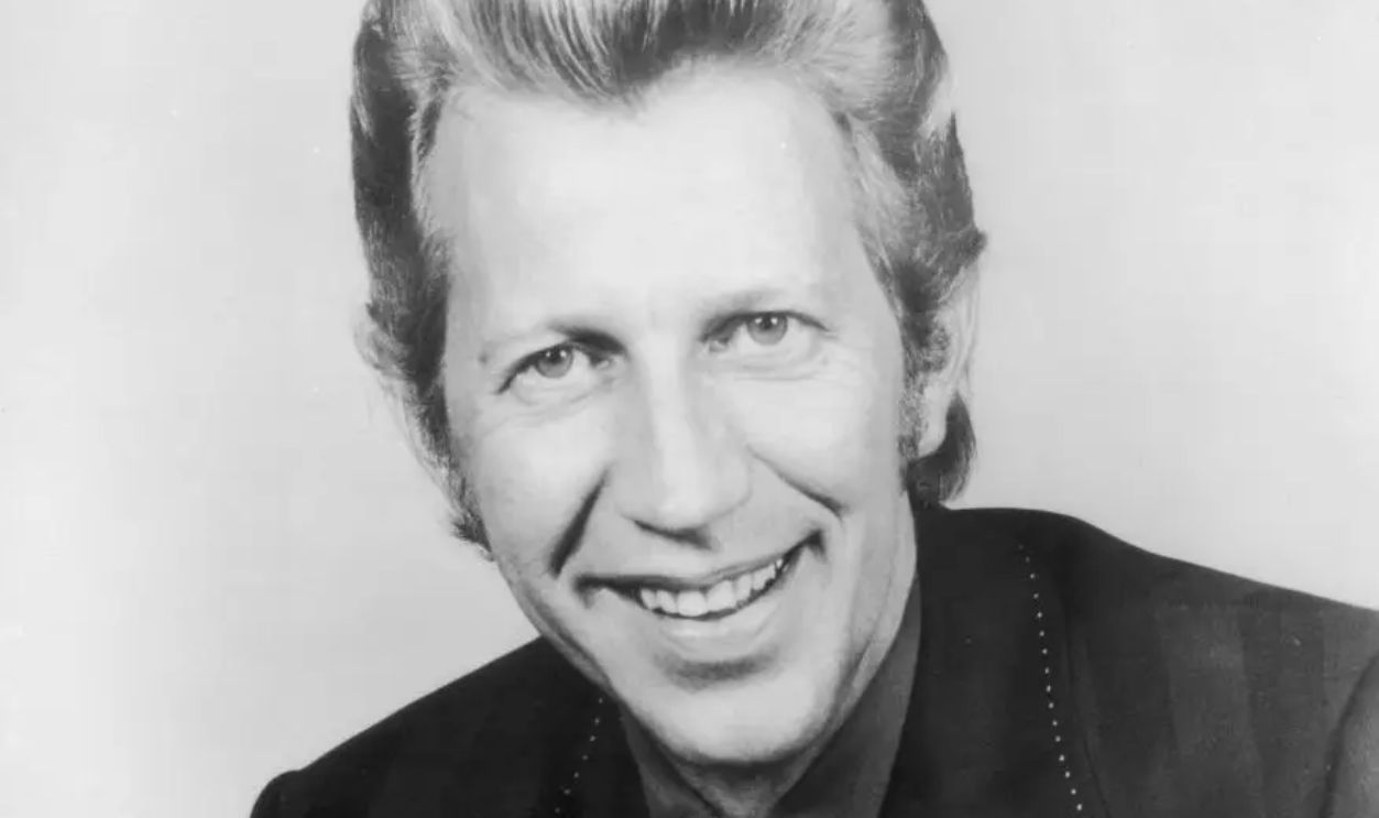 Studio portrait of American country singer Porter Wagoner smiling in a ribbon tie and a jacket with wide lapels, 1970s.