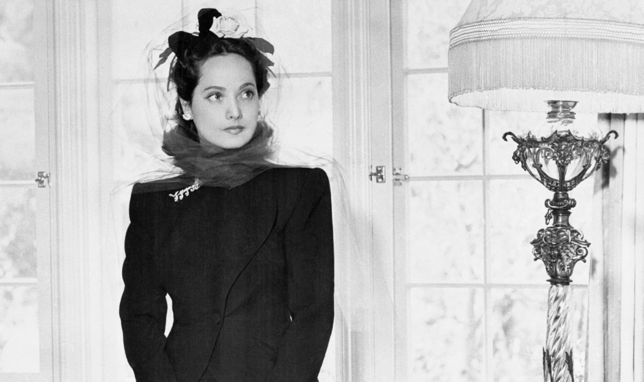 Lovely Merle Oberon poses in the library of her Bel Air home wearing a smart costume of black silk-wool.