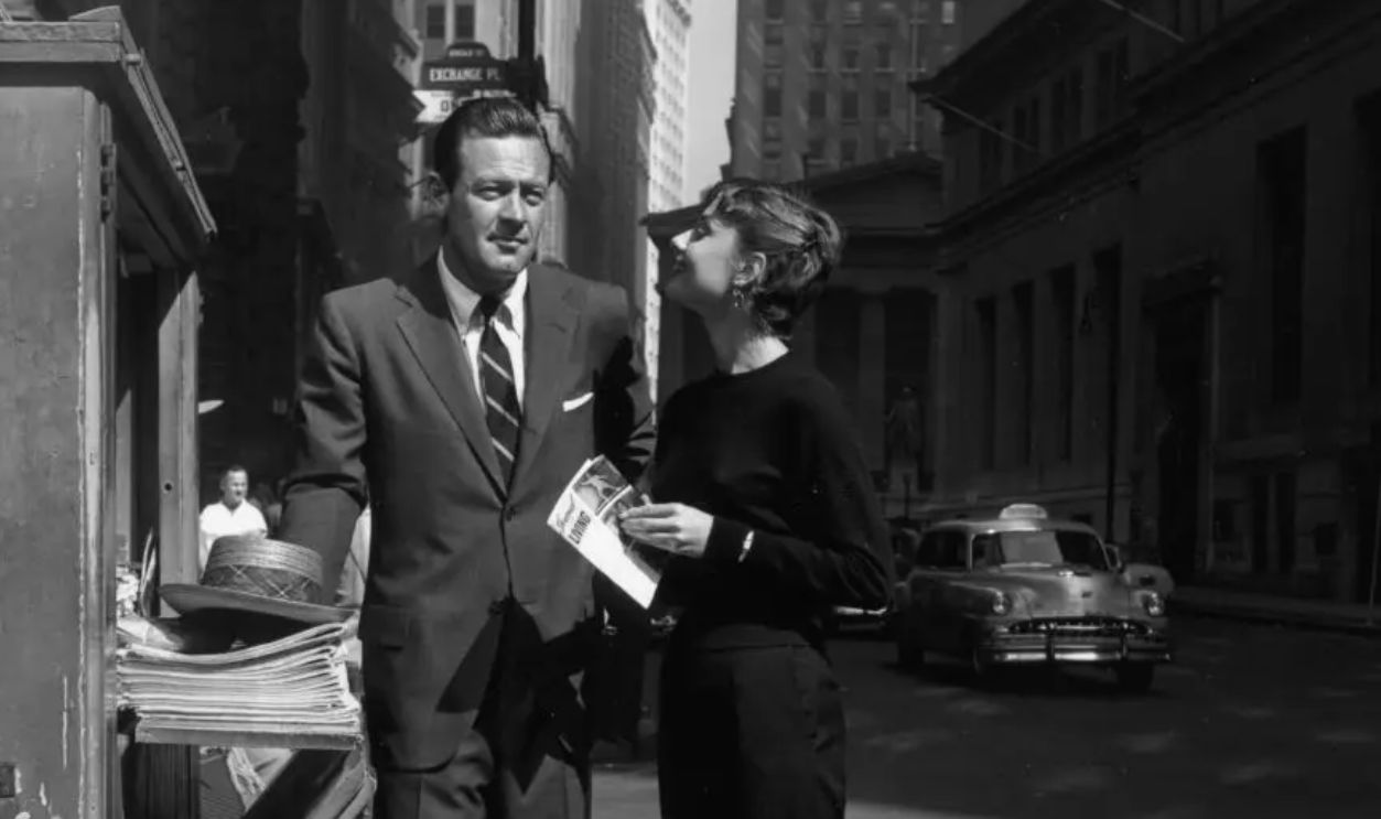 October 1953: Full-length image of American actor William Holden (1918-1981) and Belgian-born actor Audrey Hepburn (1929-1993) standing in front of a newspaper kiosk on the set of director Billy Wilder's film, 'Sabrina,' near the corner of Exchange Place, in the Wall Street district of New York City. Holden wears a suit with a rep tie. Hepburn wears a black sweater, black Capri pants, hoop earrings, and flats.