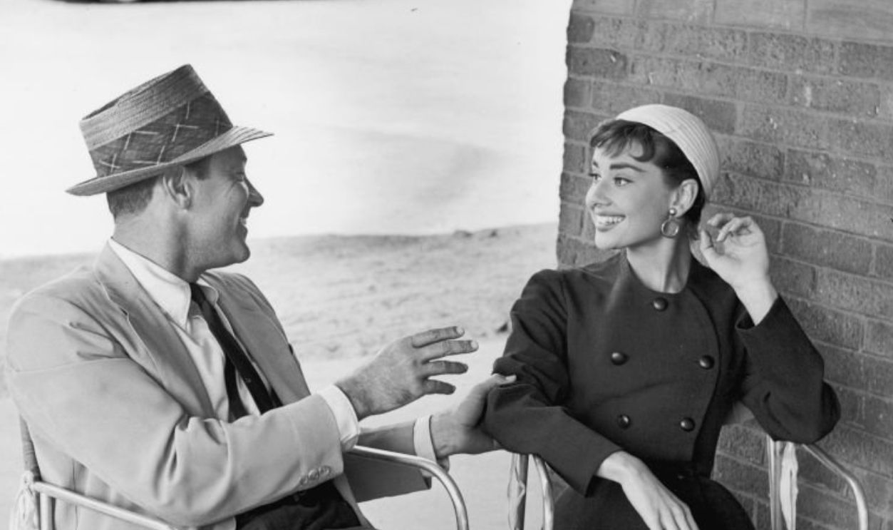 October 1953: American actor William Holden (1918 - 1981) and Belgian-born actor Audrey Hepburn (1929 - 1993) sit next to each other in chairs talking, on the set of director Billy Wilder's film, 'Sabrina'. Both are wearing hats and smiling. 
