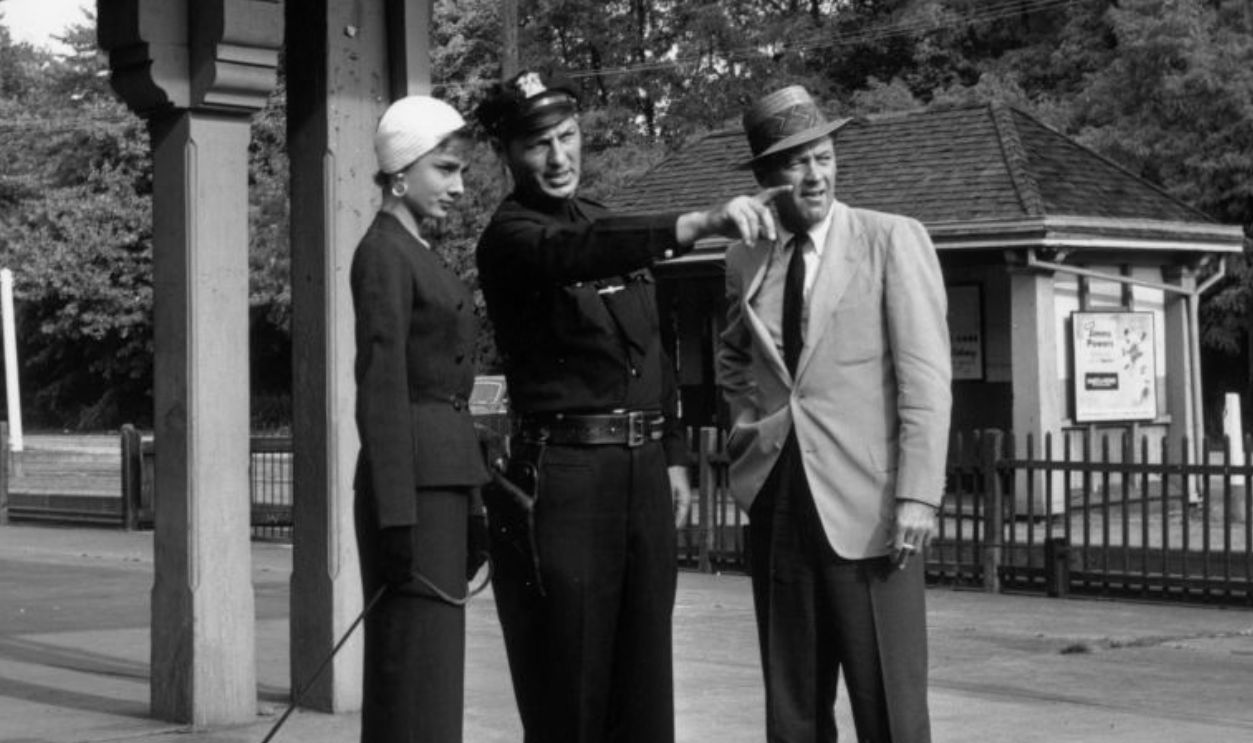 October 1953: Full-length image of Belgian-born actor Audrey Hepburn (1929-1993), holding a poodle on a leash, and American actor William Holden (1918-1981), taking directions from a police officer at a train station, on the set of director Billy Wilder's film, 'Sabrina'. Hepburn wears a skirt suit designed by Hubert de Givenchy with a hat and hoop earrings. Holden wears a straw hat and a blazer.