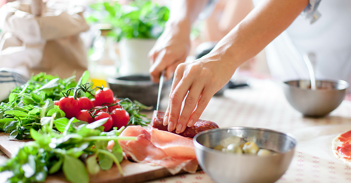 Woman is cooking in home kitchen. Female hands cut salami, vegetables, greens, tomatoes on table on wooden boards. Ingredients for preparing italian or french food. Lifestyle moment.
