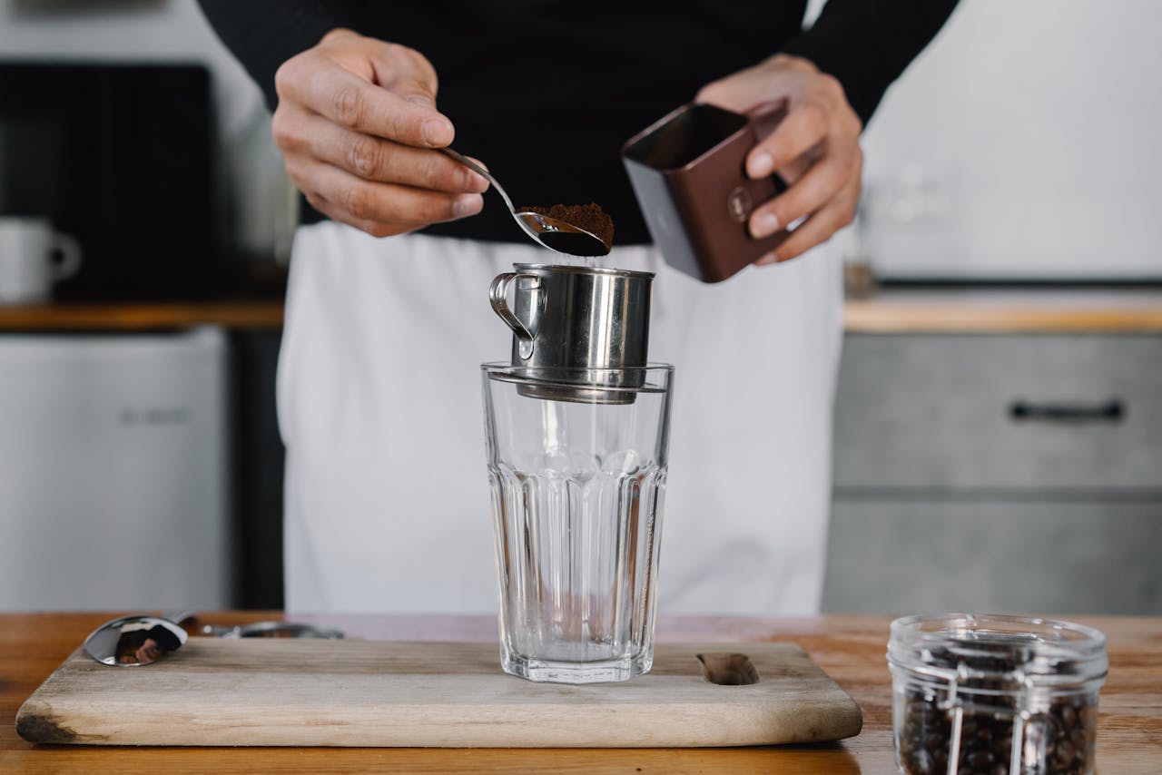 Person Putting a Scoop of Ground Coffee on a Metal Cup