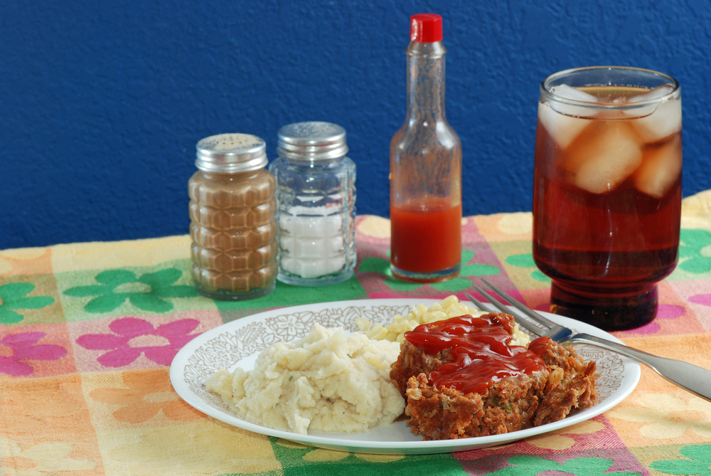 Meatloaf dinner on colorful place mat with hot sauce and salt and pepper shakers in background and served with a glass of ice tea.