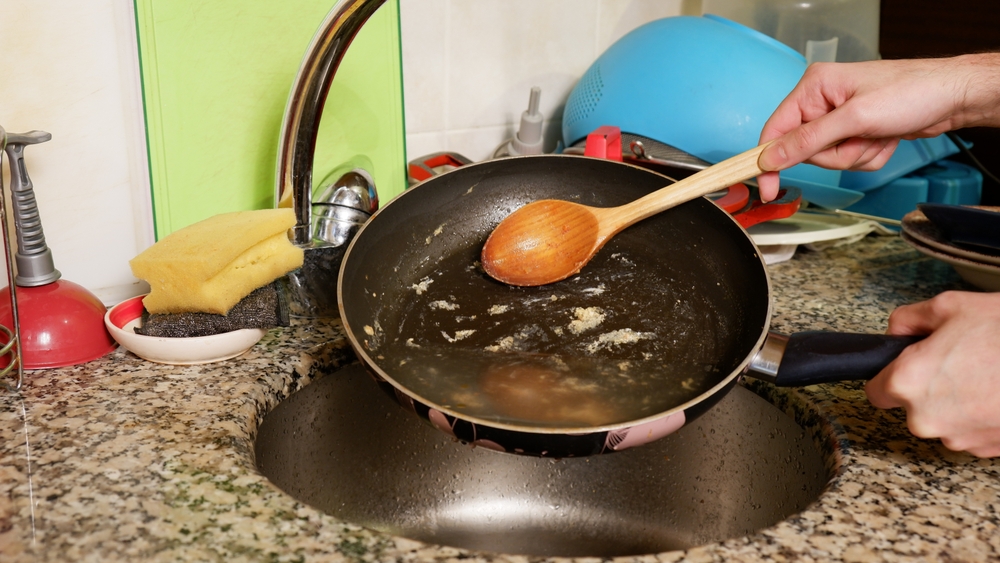 In the messy kitchen, at the sink, a man holds in one hand a dirty, greasy frying pan with watery