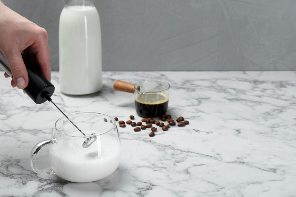 Woman whisking milk in cup with mini mixer (milk frother) at white marble table, closeup.