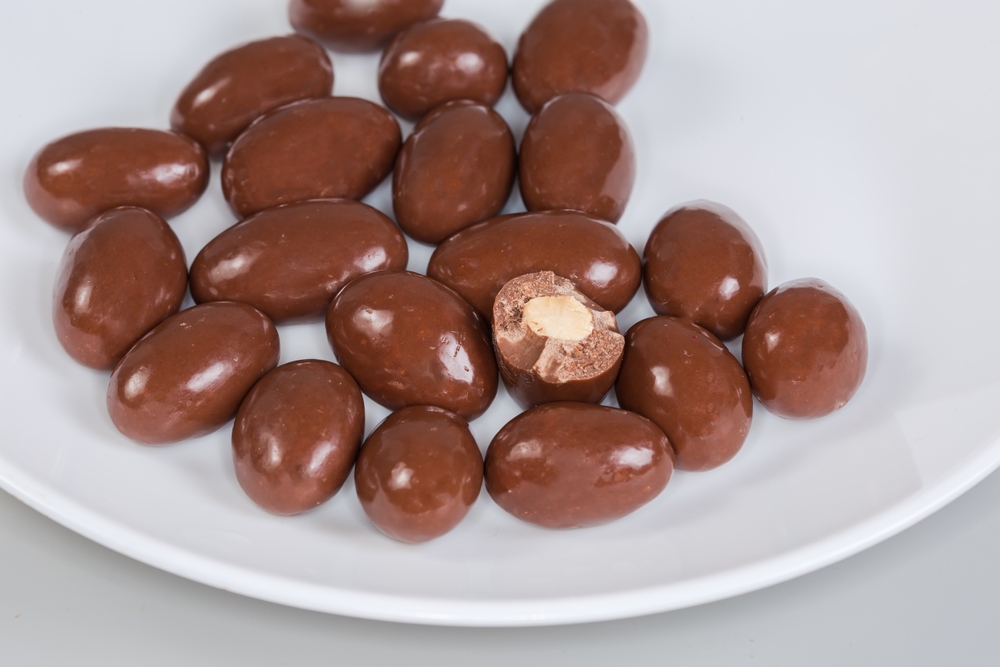 Almond nuts covered with milk chocolate on the white dish on a gray background.