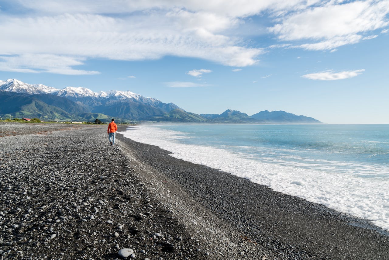 Photo of a Man in a red jacket and blue jeans Walking on Seashore Under Blue and White Sky