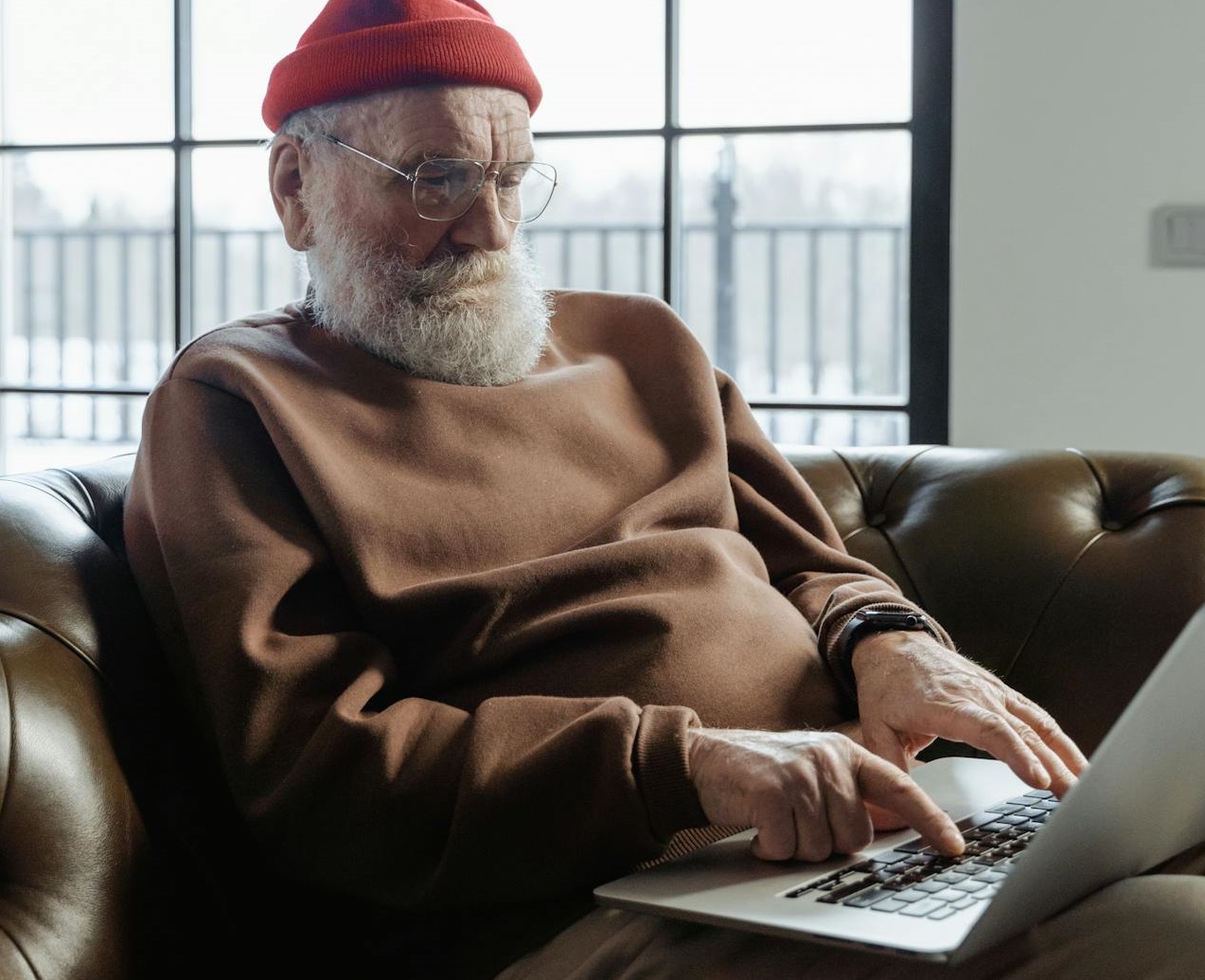 Senior man is sitting on couch and using a laptop.