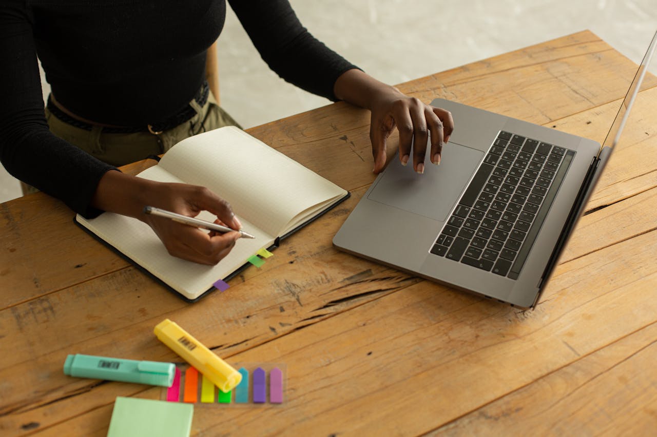Crop unrecognizable woman using laptop and writing notes while sitting on a wooden desk