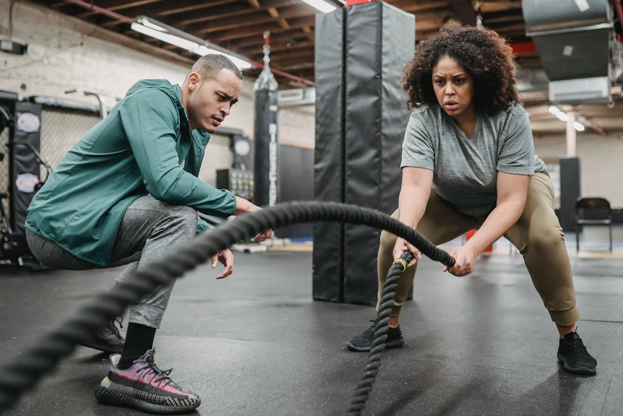 Focused black woman exercising with battling ropes near coach.