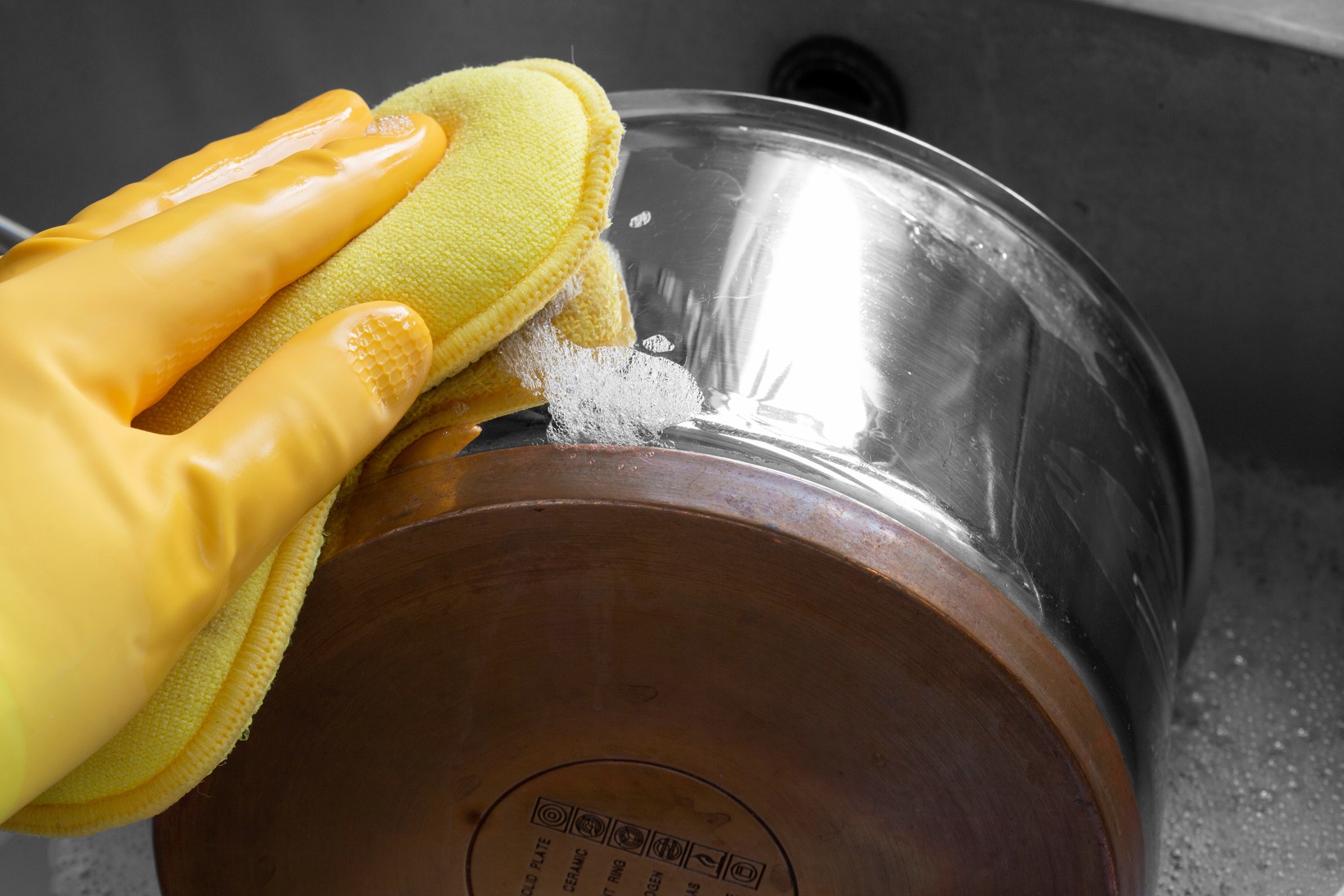 Person, wearing rubber gloves, cleaning a stainless steel and copper saucepan with a wadhing up foam pad