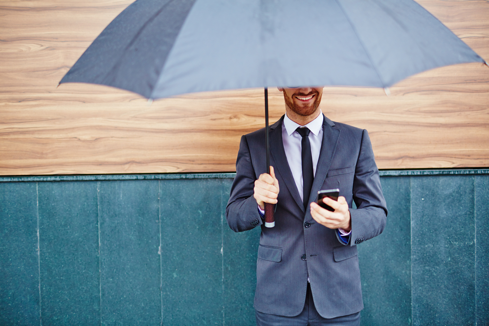Happy young businessman with smartphone standing under umbrella