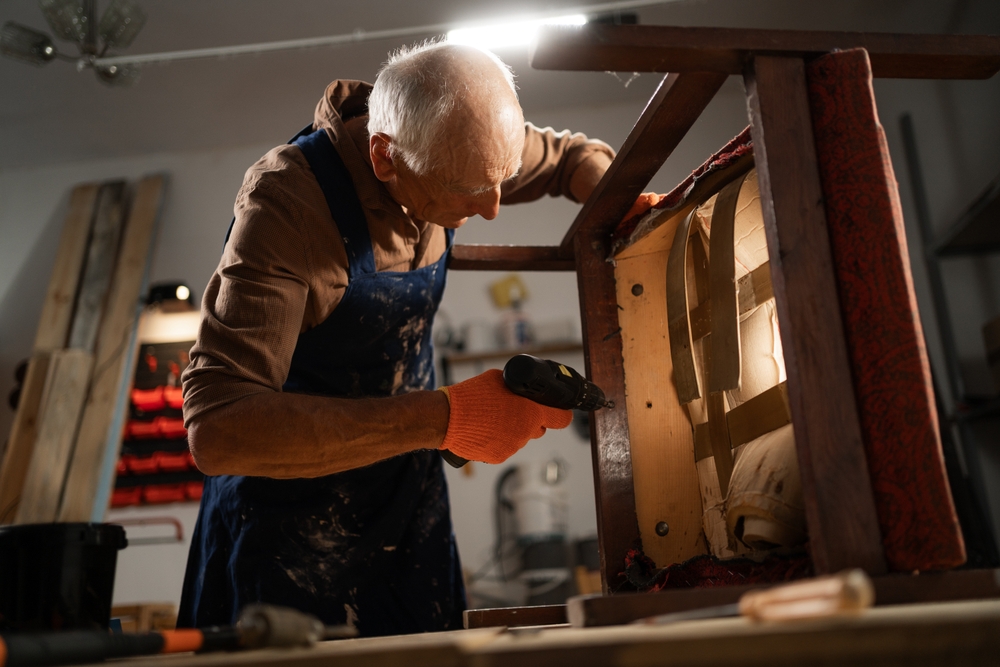An elderly man in orange gloves and a blue apron repairs a chair with an electric drill in a workshop.