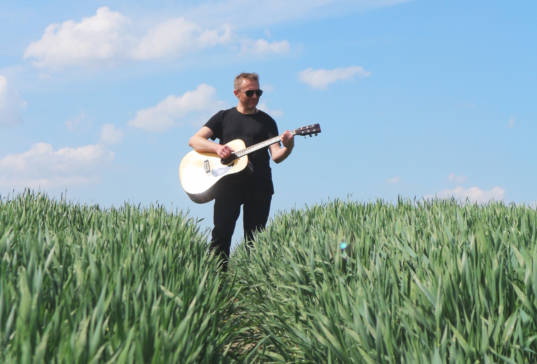 A man in black sunglasses with a guitar in the field.