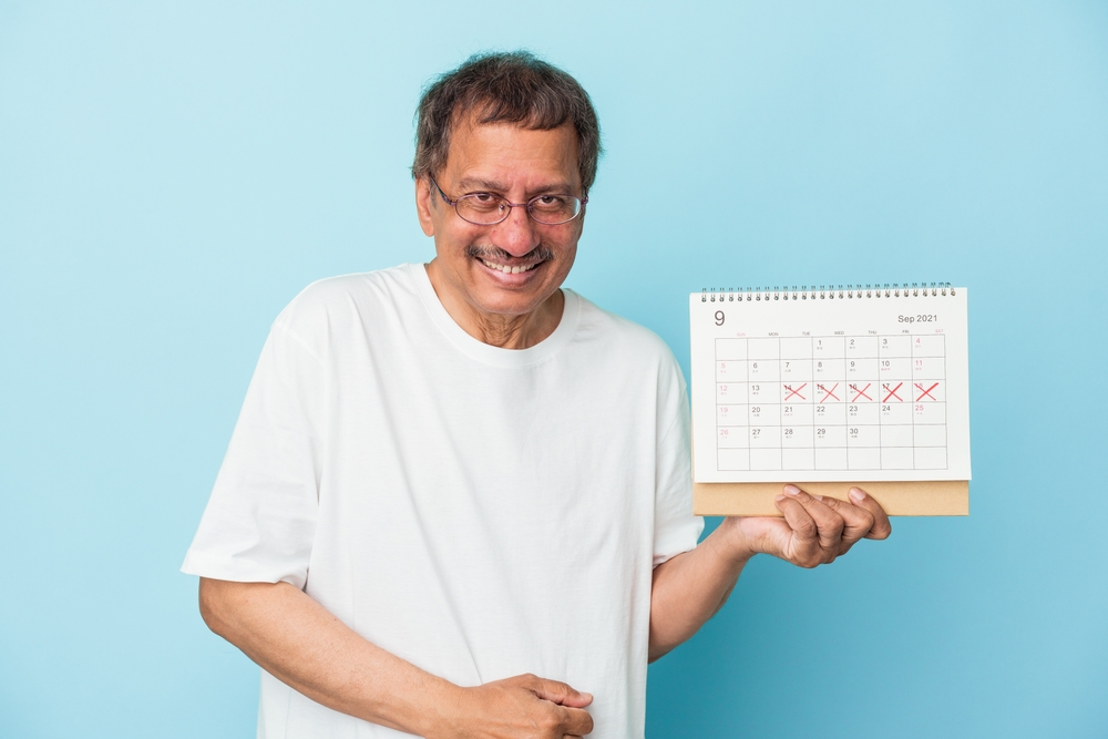 Senior indian man holding a calendar isolated on blue background laughing and having fun.