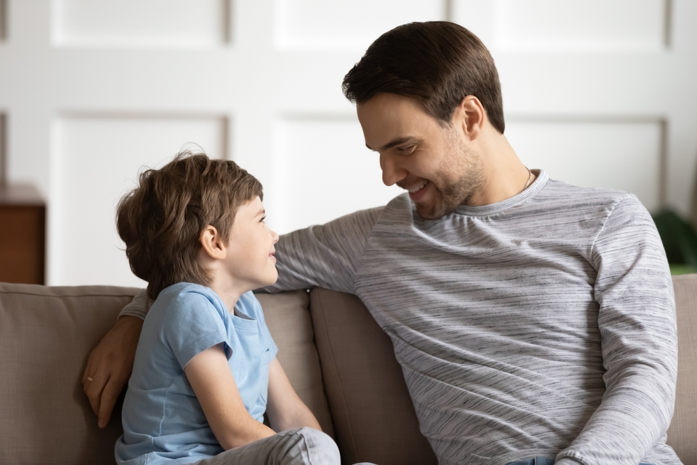 Happy young father and little son sit on couch in living room talk chat on leisure family weekend.