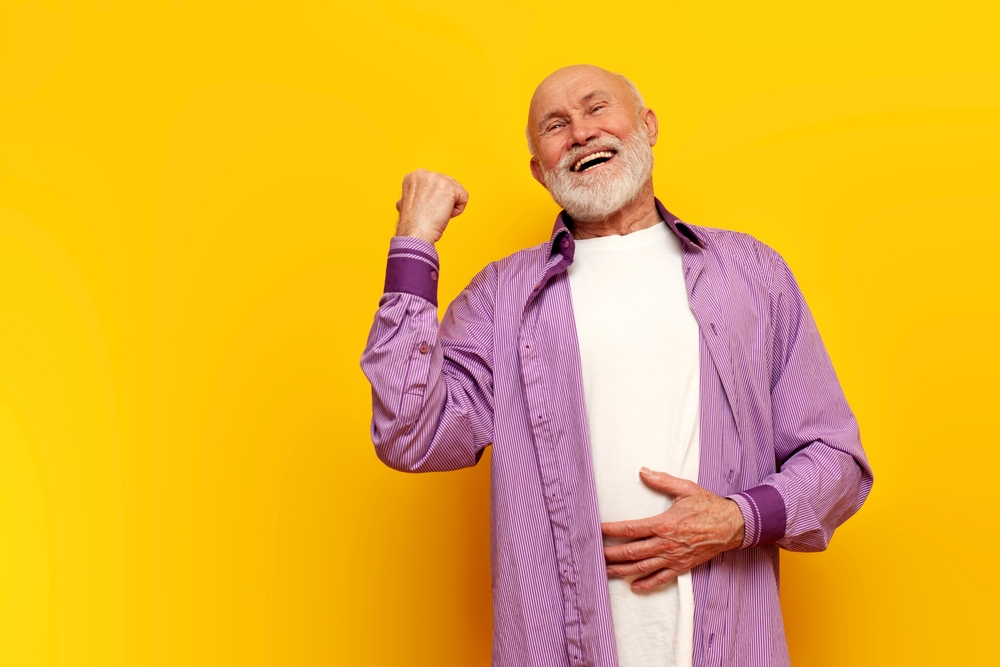 old bald grandfather with gray beard points to the side and laughs on yellow isolated background
