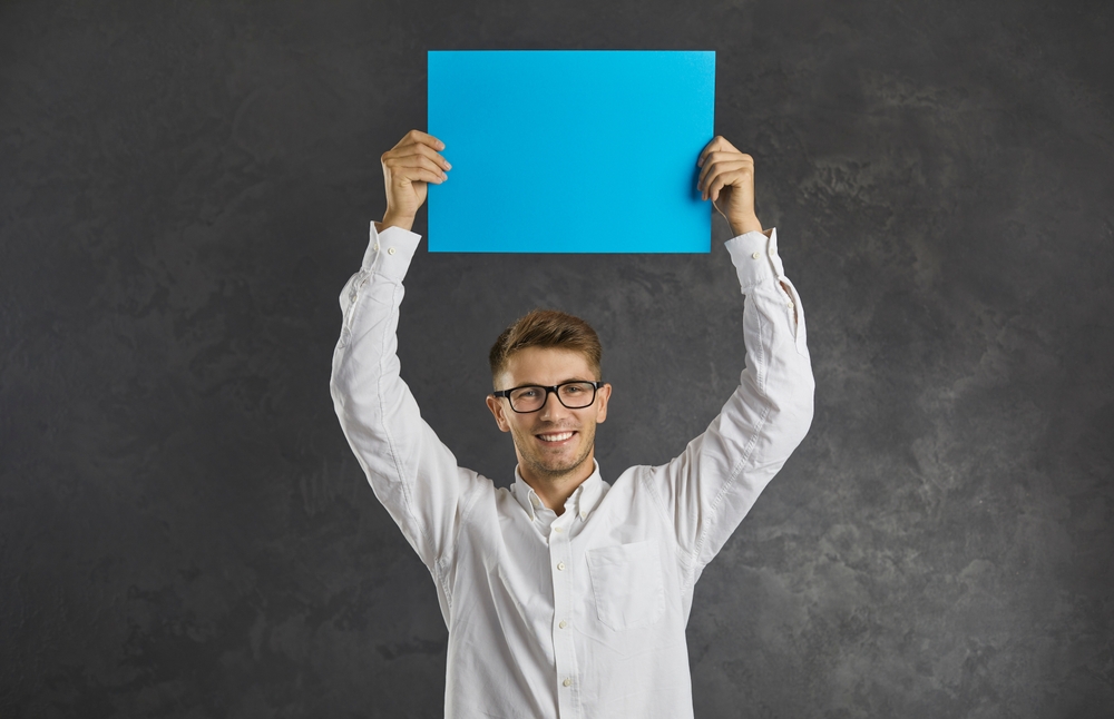 Portrait of smiling millennial gen z man isolated on black studio background hold blue mockup paper.