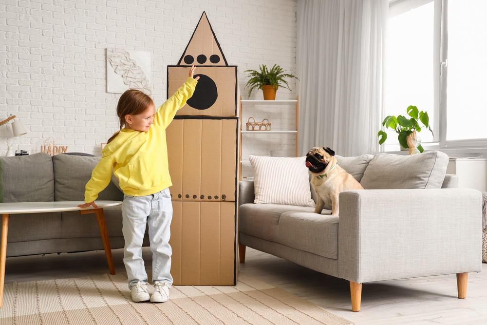 Little girl playing with cardboard rocket and pug dog at home
